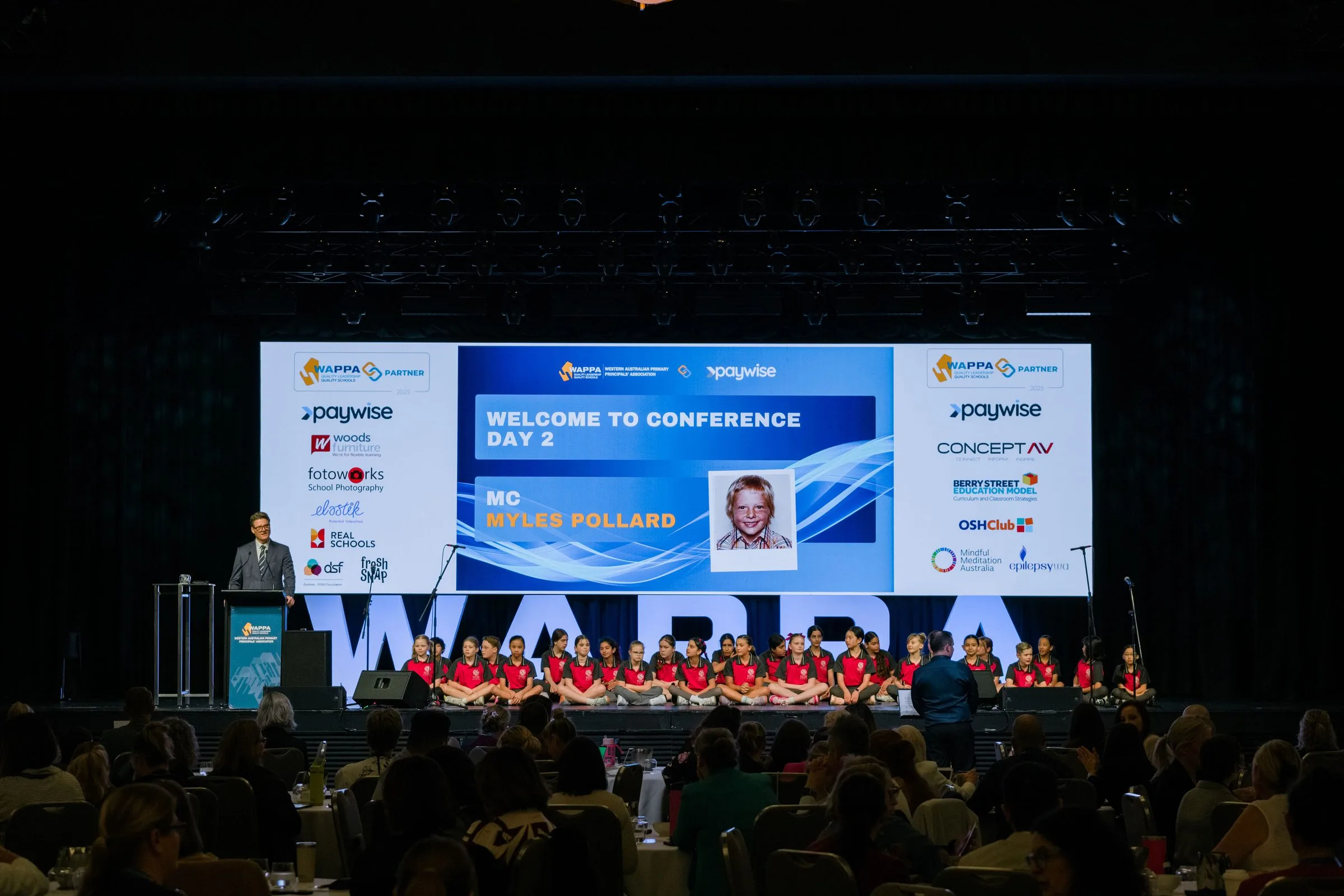 Conference stage screens welcoming guests to day two, MC Myles Pollard
