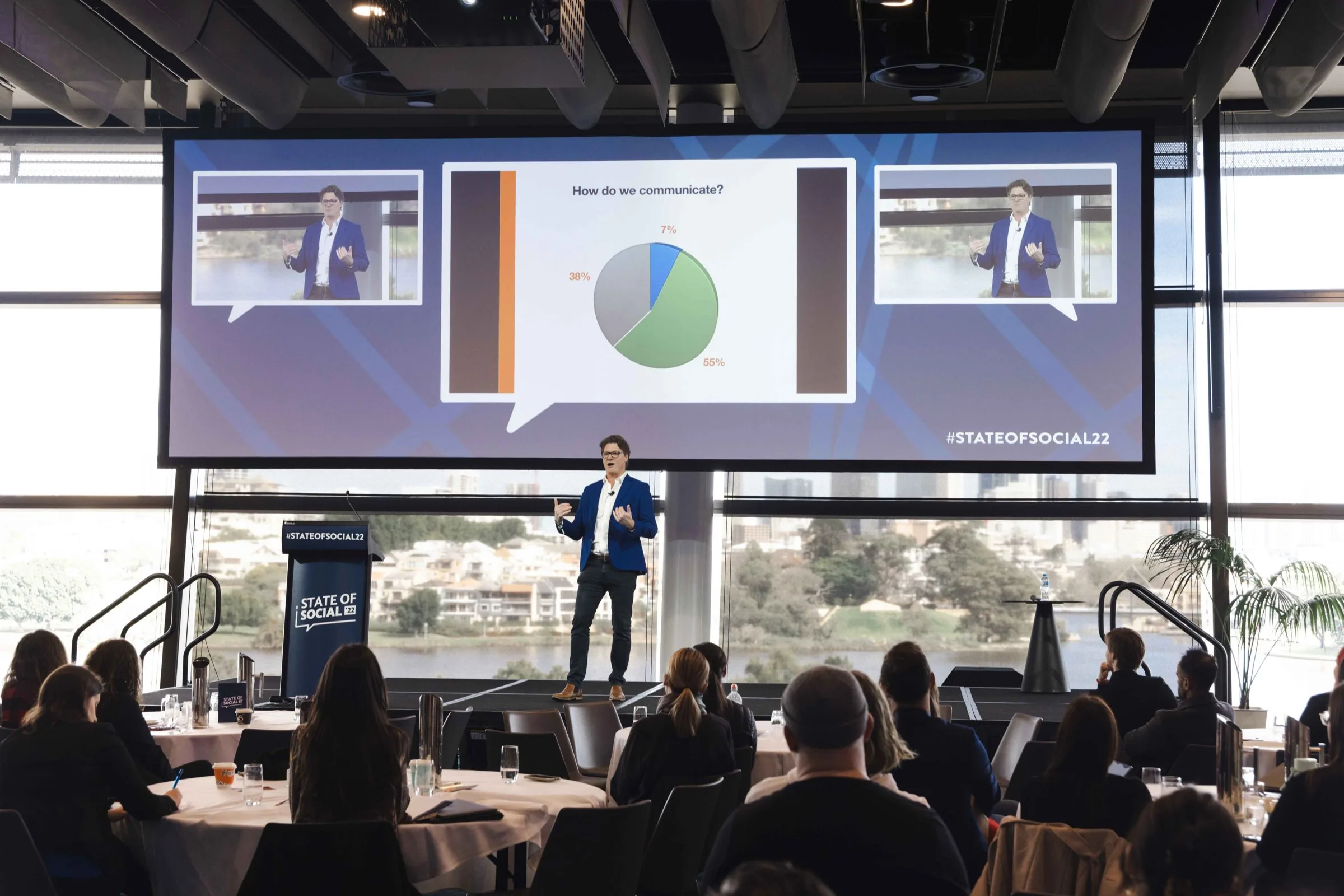 Speaker presenting to a conference audience beside a large presentation screen